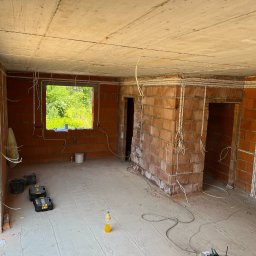 PS Energia Piotr Sitnik - Wnętrze w stanie surowym z widoczną instalacją elektryczną w peszlach, prowadzoną po ścianach i suficie, z narzędziami i butelką na podłodze.