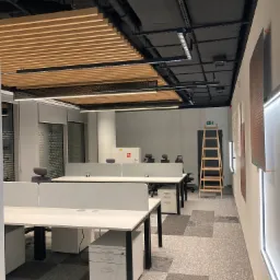 Nowoczesne biuro z zainstalowanym oświetleniem podsufitowym, widoczne biurka z szarymi przegrodami, drabina oparta o ścianę w tle oraz czarna instalacja elektryczna na suficie.