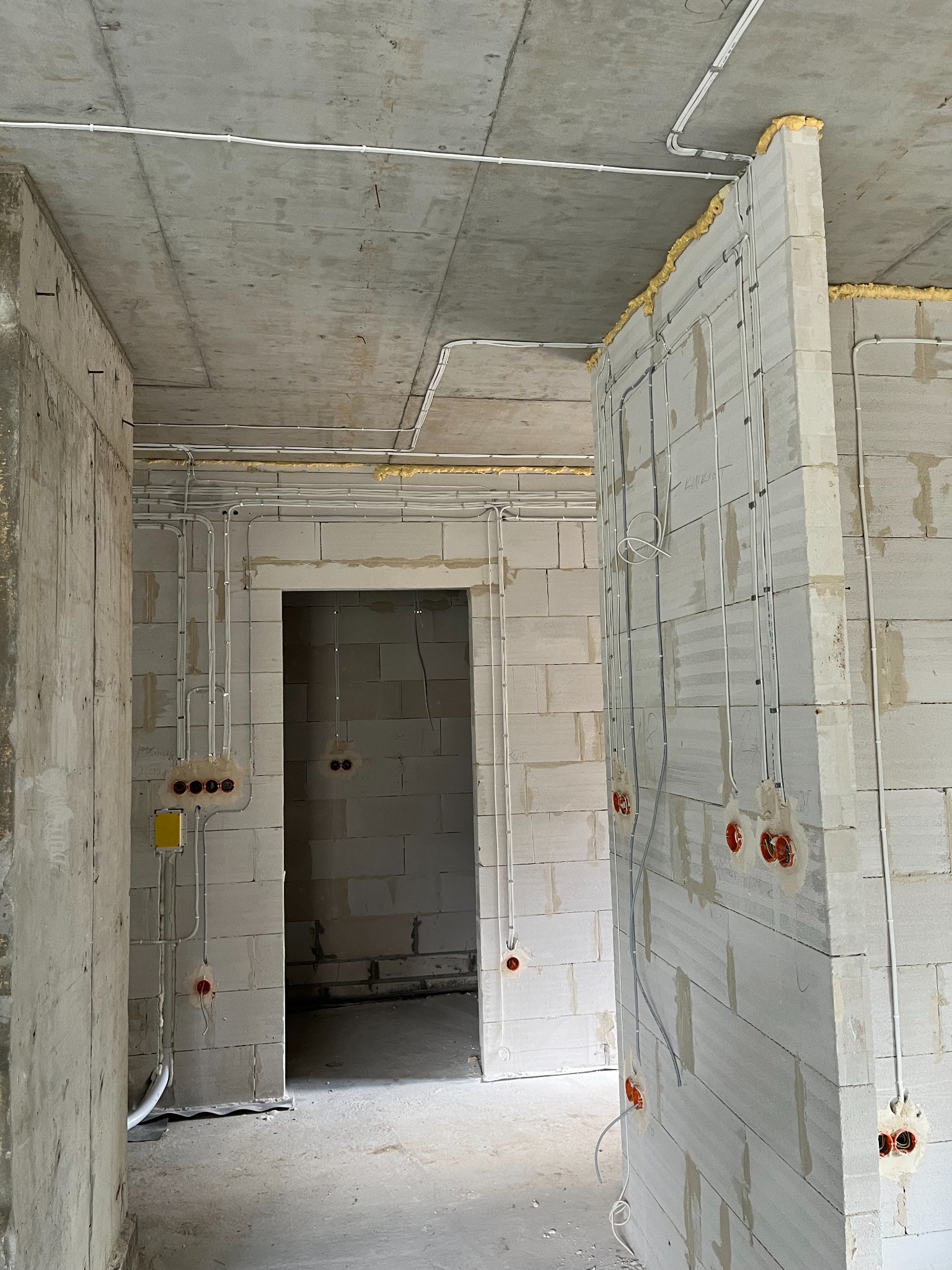 Wnętrze w stanie surowym z widocznymi przewodami elektrycznymi poprowadzonymi po ścianach z betonu komórkowego oraz pod sufitem, zabezpieczonymi pianką montażową w narożnikach.