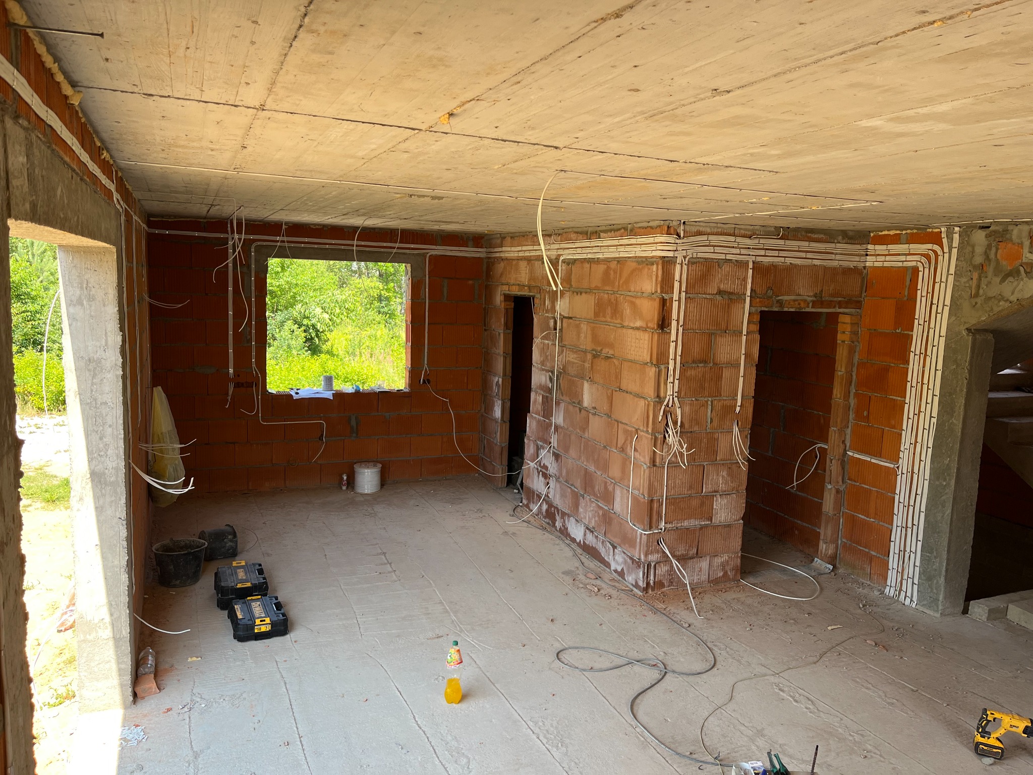 Wnętrze w stanie surowym z widoczną instalacją elektryczną w peszlach, prowadzoną po ścianach i suficie, z narzędziami i butelką na podłodze.