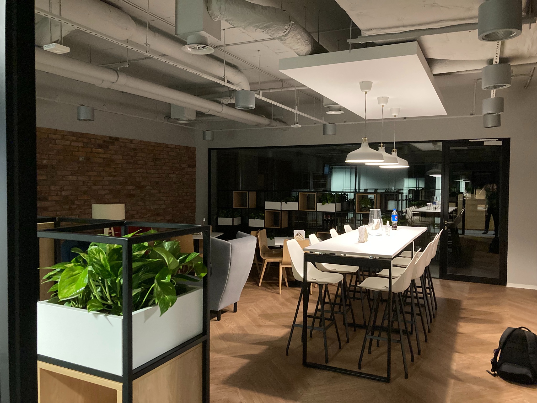 Oświetlenie biurowe z widocznymi instalacjami pod sufitem w Maciejowicach, z lampami wiszącymi nad stołem i widocznymi przewodami oraz elementami wentylacji.