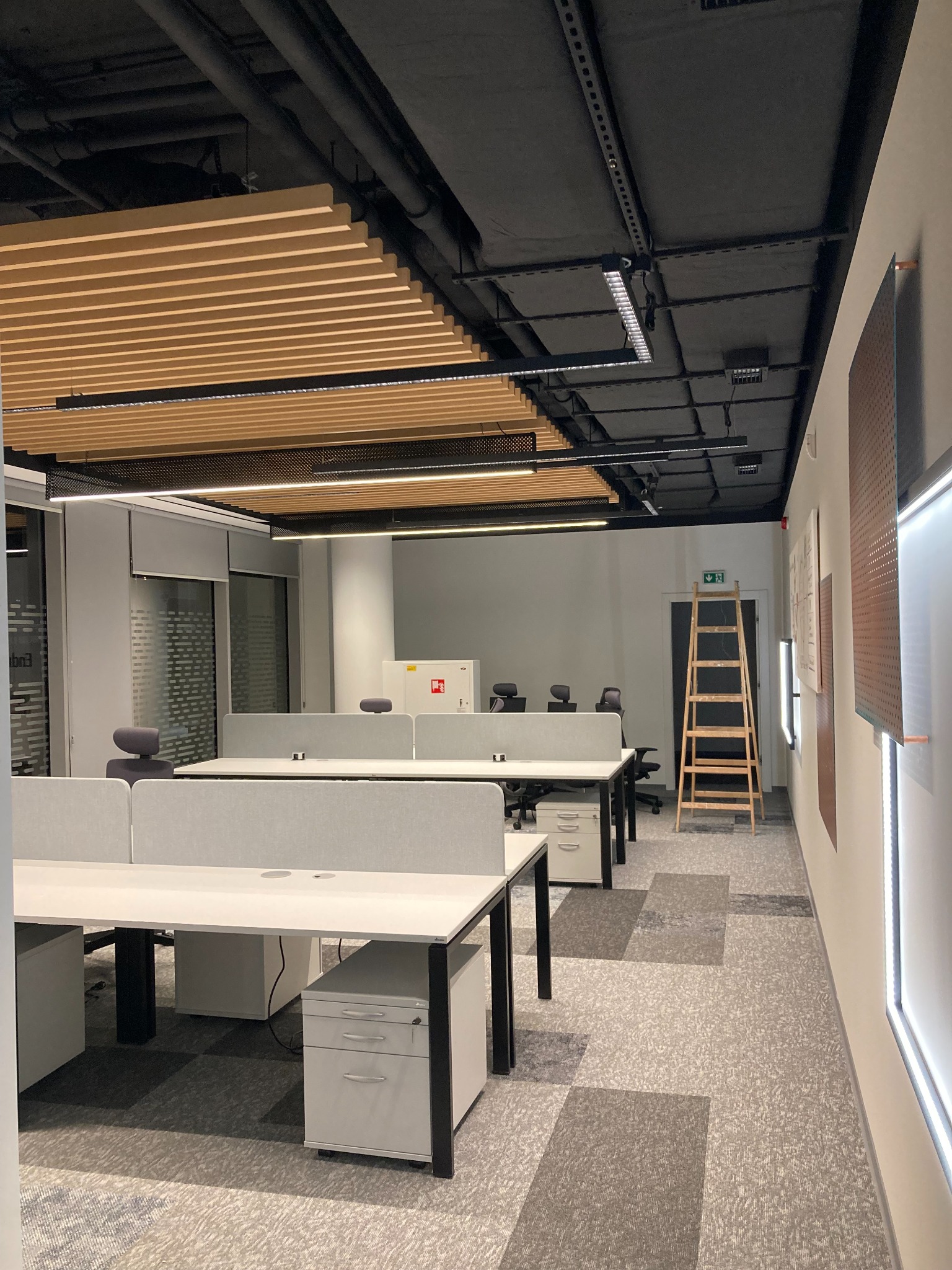 Nowoczesne biuro z zainstalowanym oświetleniem podsufitowym, widoczne biurka z szarymi przegrodami, drabina oparta o ścianę w tle oraz czarna instalacja elektryczna na suficie.