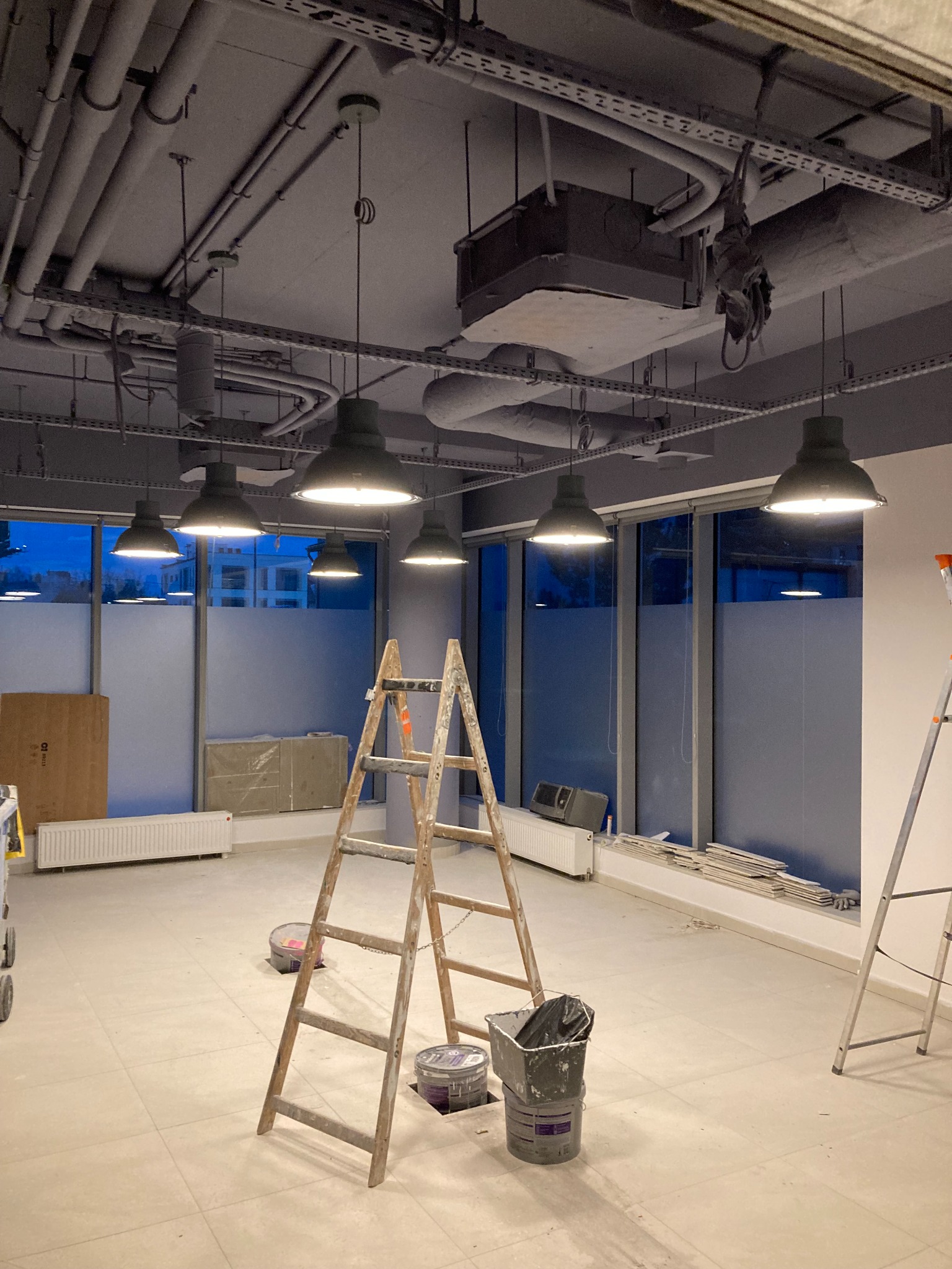 Wnętrze biurowe w trakcie wykańczania, widoczne instalacje pod sufitem, lampy przemysłowe, drabiny malarskie i puszki z farbą.
