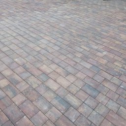 HYDRO-MAR SUSŁYK - Ułożona, prostokątna kostka brukowa w odcieniach brązu i szarości, tworząca wzór na podjeździe. W tle widać stosy kostki przygotowane do dalszego układania.