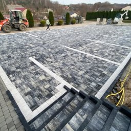 HYDRO-MAR SUSŁYK - Ukończona nawierzchnia z kostki brukowej w szaro-białej kolorystyce, tworząca wzór parkingu z wyznaczonymi miejscami. Widoczny fragment schodów z kostki i maszyny budowlane w tle.