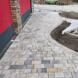 Kaskadowa ścieżka z kostki brukowej w odcieniach szarości i beżu, wijąca się obok budynku z ceglanym narożnikiem, z widocznym zapleczem budowy w tle.