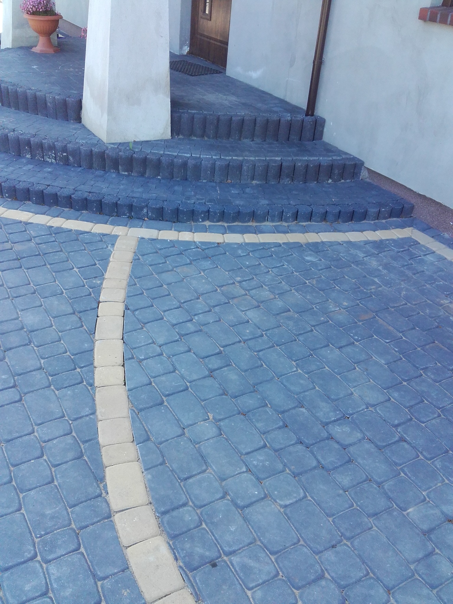 Ułożona, niebieska kostka brukowa z kremową obwódką tworzącą wzór. Schody z ciemnych, okrągłych kostek prowadzą do wejścia.