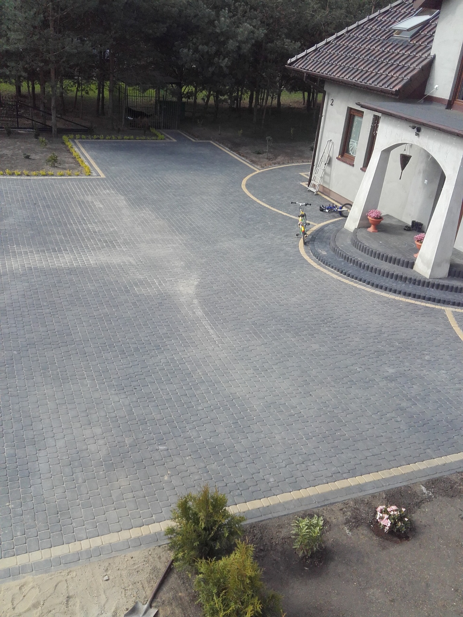 Rozległy podjazd z szarej kostki brukowej z żółtymi obrzeżami, prowadzący do domu z łukowym wejściem. Widoczne rośliny, rower i drabina oparta o ścianę.