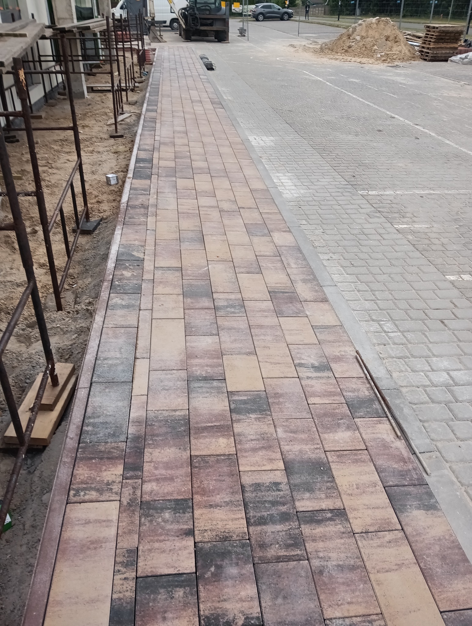 Ułożona, wielobarwna kostka brukowa na nowym chodniku. W tle widoczny mini-koparka, budynek w rusztowaniach i hałda piasku. Widok z perspektywy osoby stojącej.