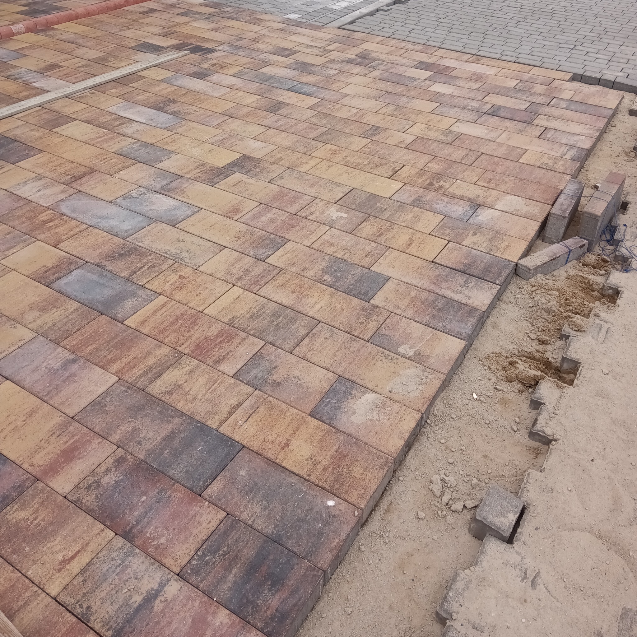 Układanie prostokątnej kostki brukowej w odcieniach brązu i szarości, widoczne niedokończone krawędzie i przygotowane podłoże z piasku.