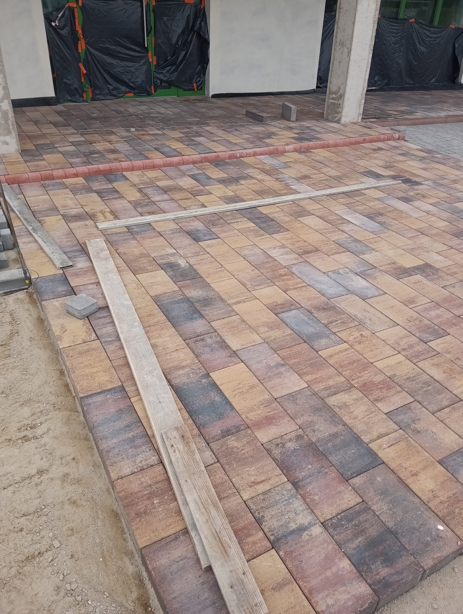 Częściowo ułożona, wielobarwna kostka brukowa na podwórku. Widoczne drewniane listwy i pojedyncze kostki, sugerujące trwające prace. Tło z czarną folią ochronną.