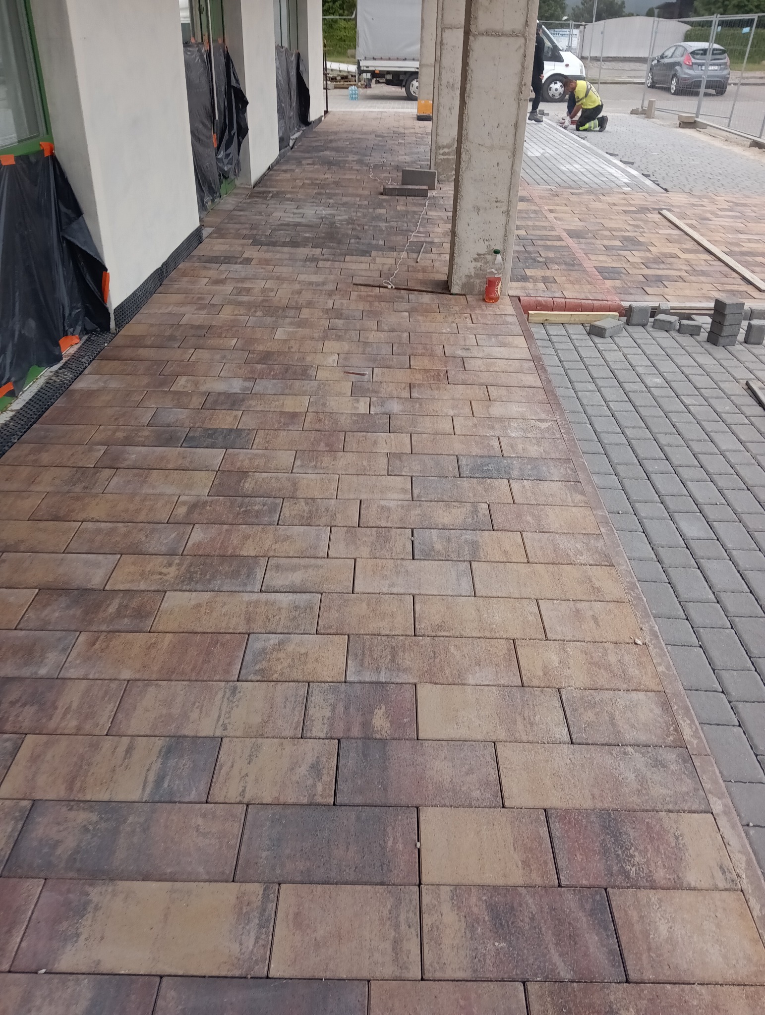 Fragment układania kostki brukowej w odcieniach brązu i szarości przy budynku. Widoczny pracownik w tle, przygotowane kostki i betonowe słupy.