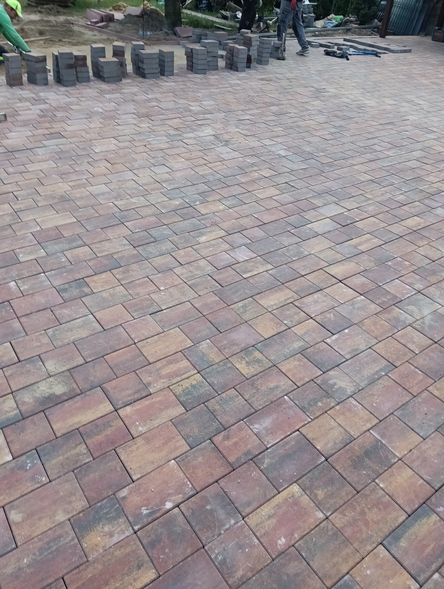 Fragment nowej nawierzchni z kostki brukowej w odcieniach brązu i szarości, z widocznymi narzędziami brukarskimi i stosem kostek w tle.