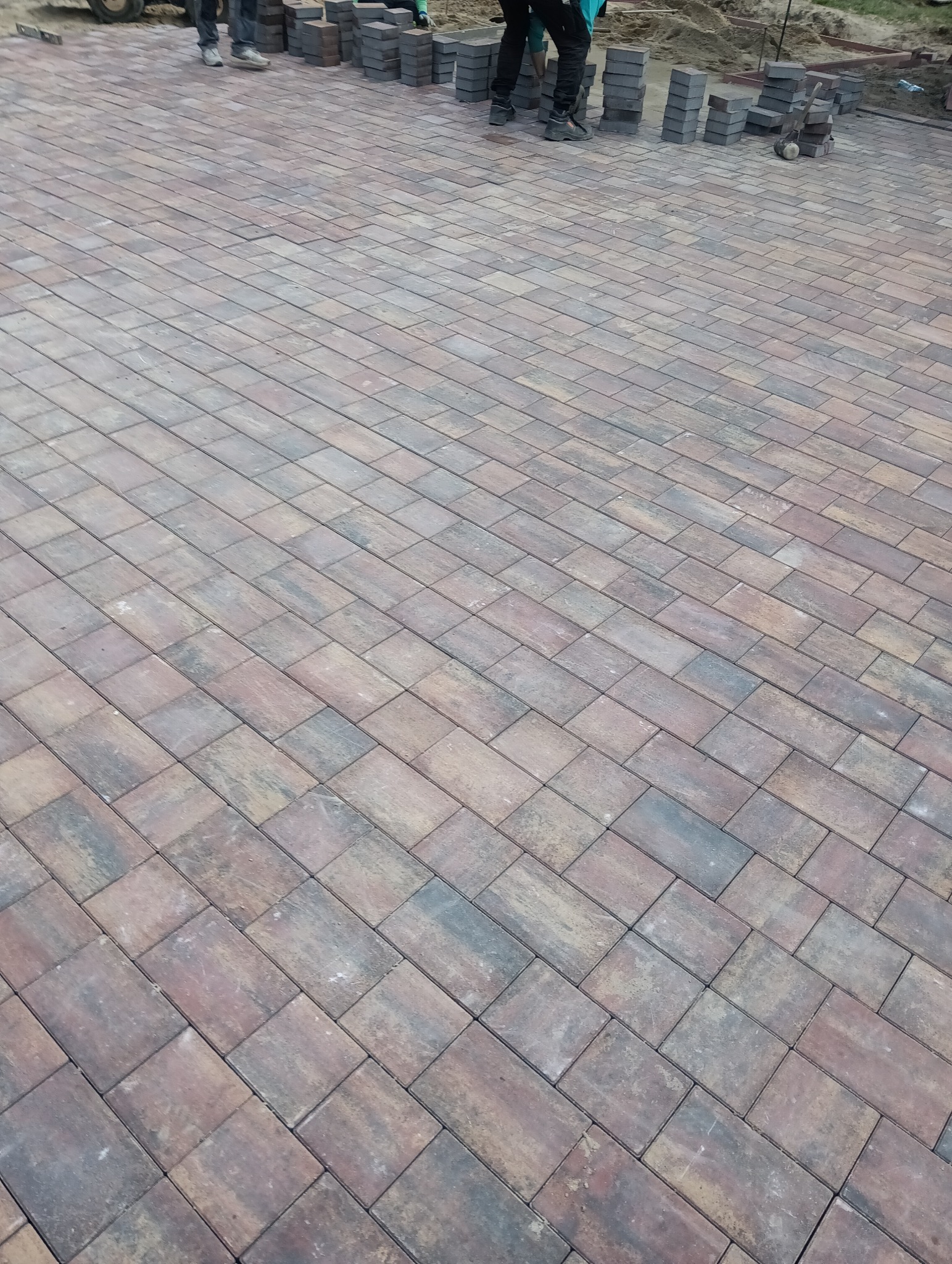 Ułożona, prostokątna kostka brukowa w odcieniach brązu i szarości, tworząca wzór na podjeździe. W tle widać stosy kostki przygotowane do dalszego układania.