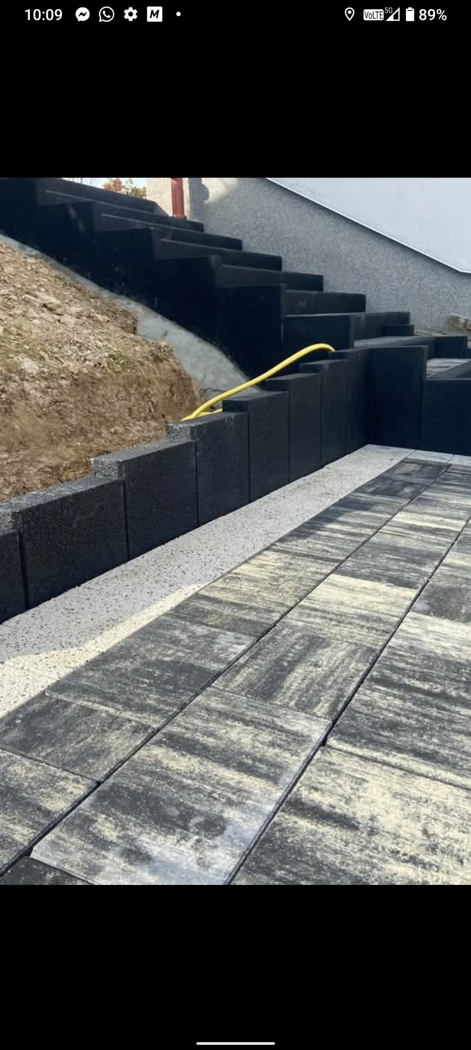 Ułożona kostka brukowa w odcieniach szarości, tworząca ścieżkę i schody. Widoczny fragment żółtego węża. Ścieżka oddzielona od murku białym żwirem.