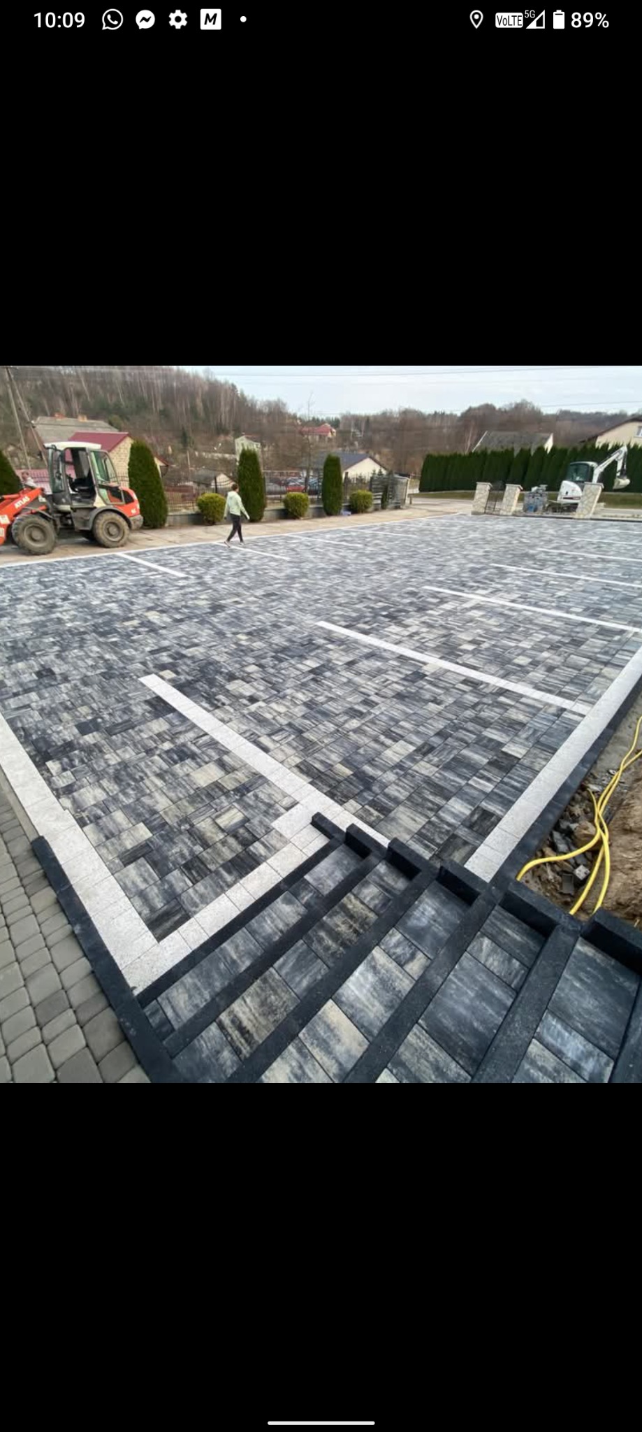 Ukończona nawierzchnia z kostki brukowej w szaro-białej kolorystyce, tworząca wzór parkingu z wyznaczonymi miejscami. Widoczny fragment schodów z kostki i maszyny budowlane w tle.