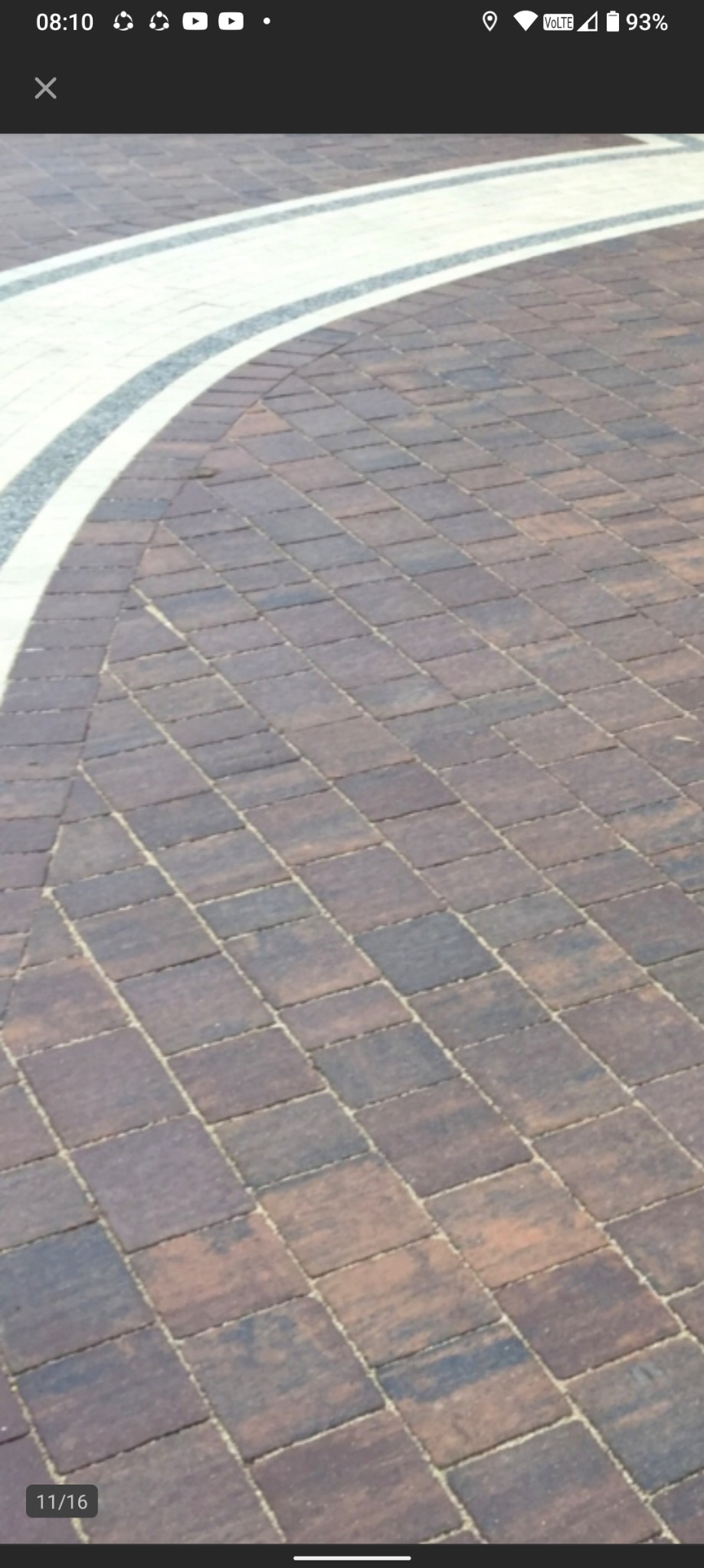 Ułożona, brązowa kostka brukowa z białym obramowaniem, tworząca zakrzywiony wzór na podjeździe. Widok z góry, ukośny kadr.