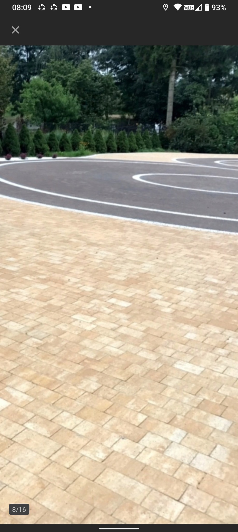 Ułożona beżowa kostka brukowa z elementami asfaltu i białymi liniami, tworząca wzór na podjeździe. W tle widoczne krzewy i drzewa.