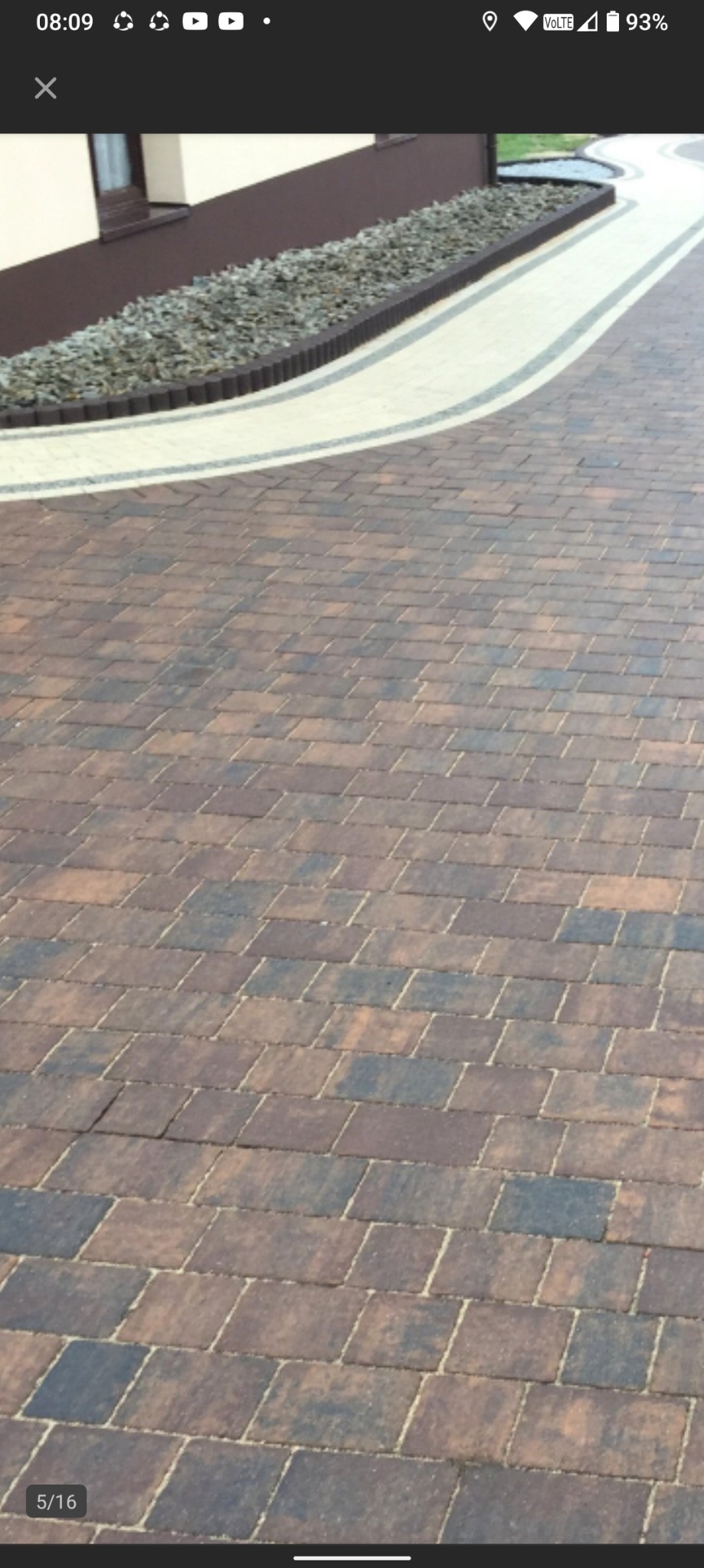 Ułożona, brązowo-szara kostka brukowa na podjeździe, z obrzeżem z jasnych kostek i rabatą z kamieni przy budynku. Widok z góry, lekko z ukosa.