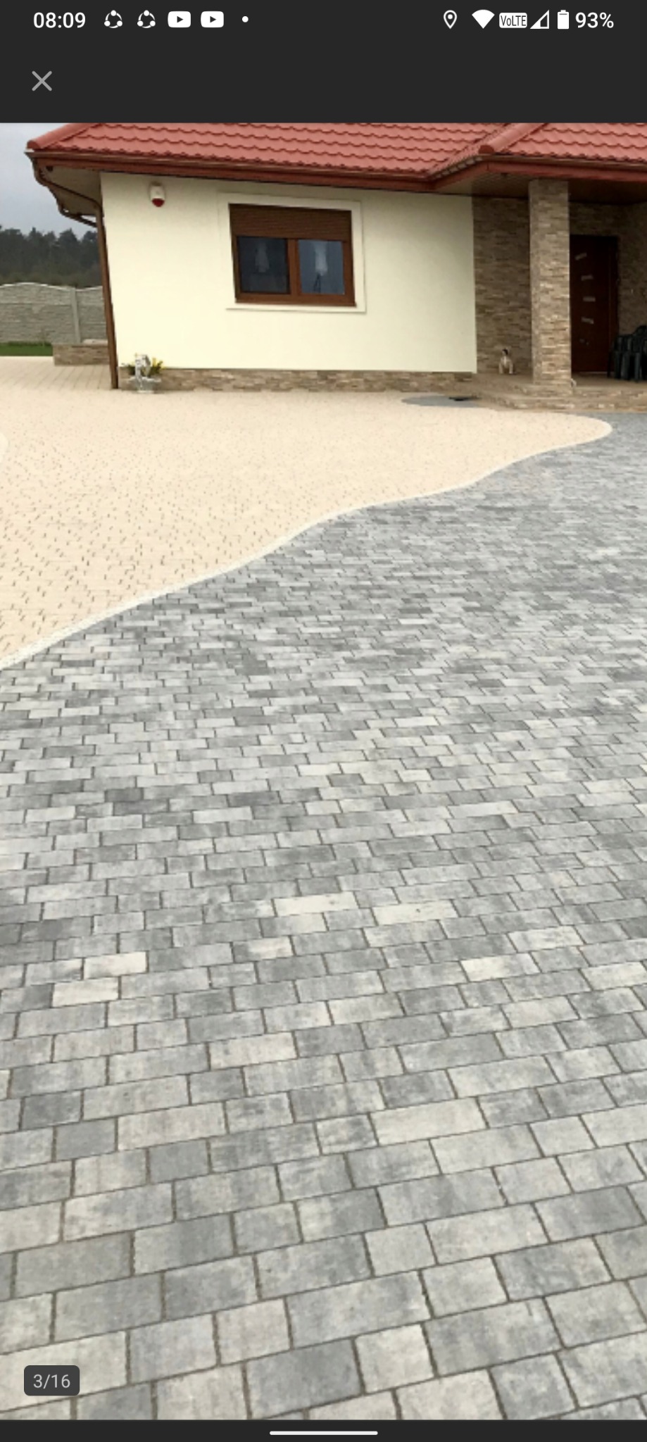 Dwukolorowy podjazd z kostki brukowej w odcieniach szarości i beżu, prowadzący do domu z brązowym dachem. Widoczny fragment elewacji i mały pies na schodach.