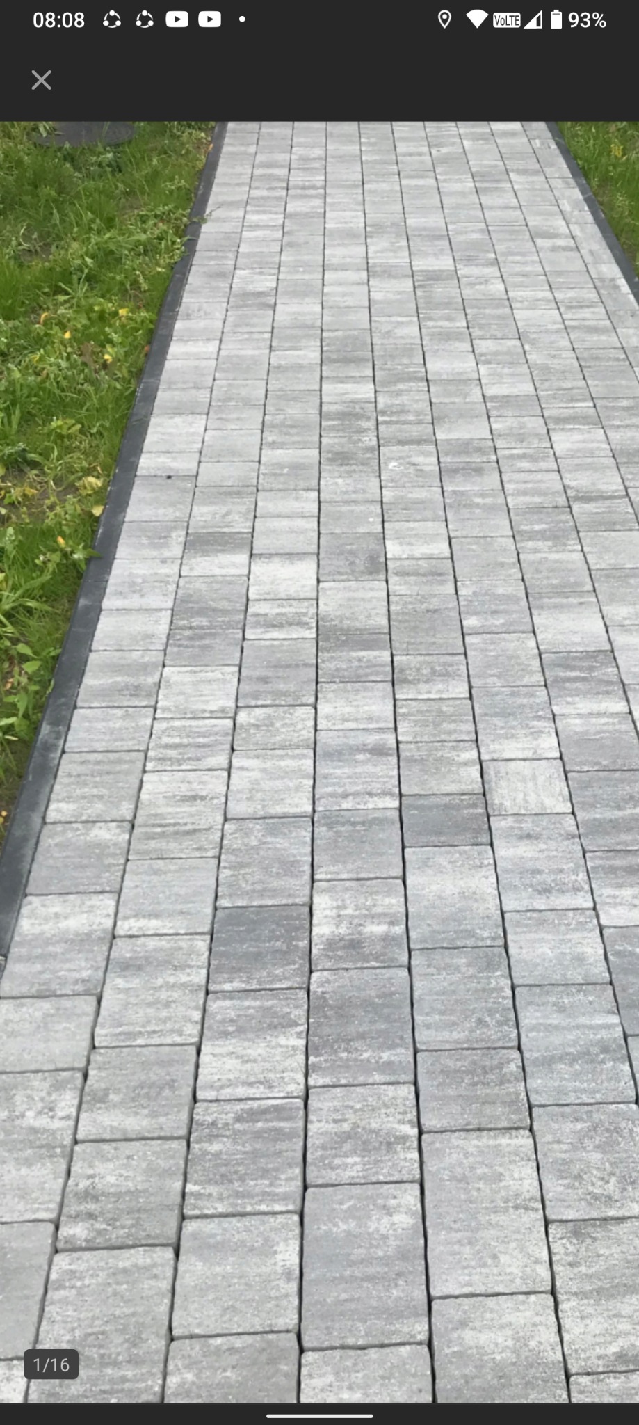 Ułożona ścieżka z szarej kostki brukowej, widok z góry, z czarnym obramowaniem po bokach i zieloną trawą w tle. Regularny wzór kostki.