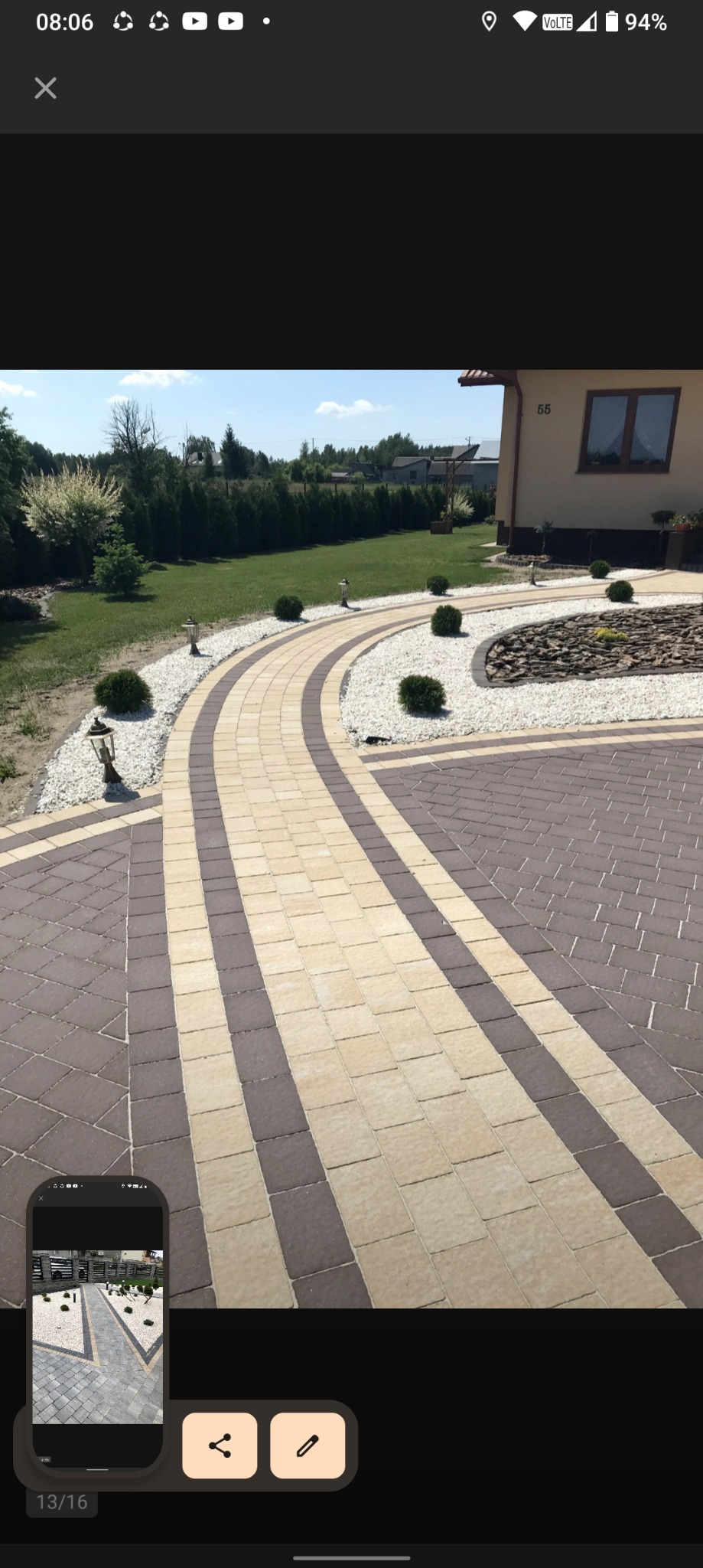 Zakrzywiony chodnik z kostki brukowej w dwóch odcieniach, obłożony białym kamieniem. Widoczne lampki ogrodowe i trawnik. Dom z numerem 55 w tle.