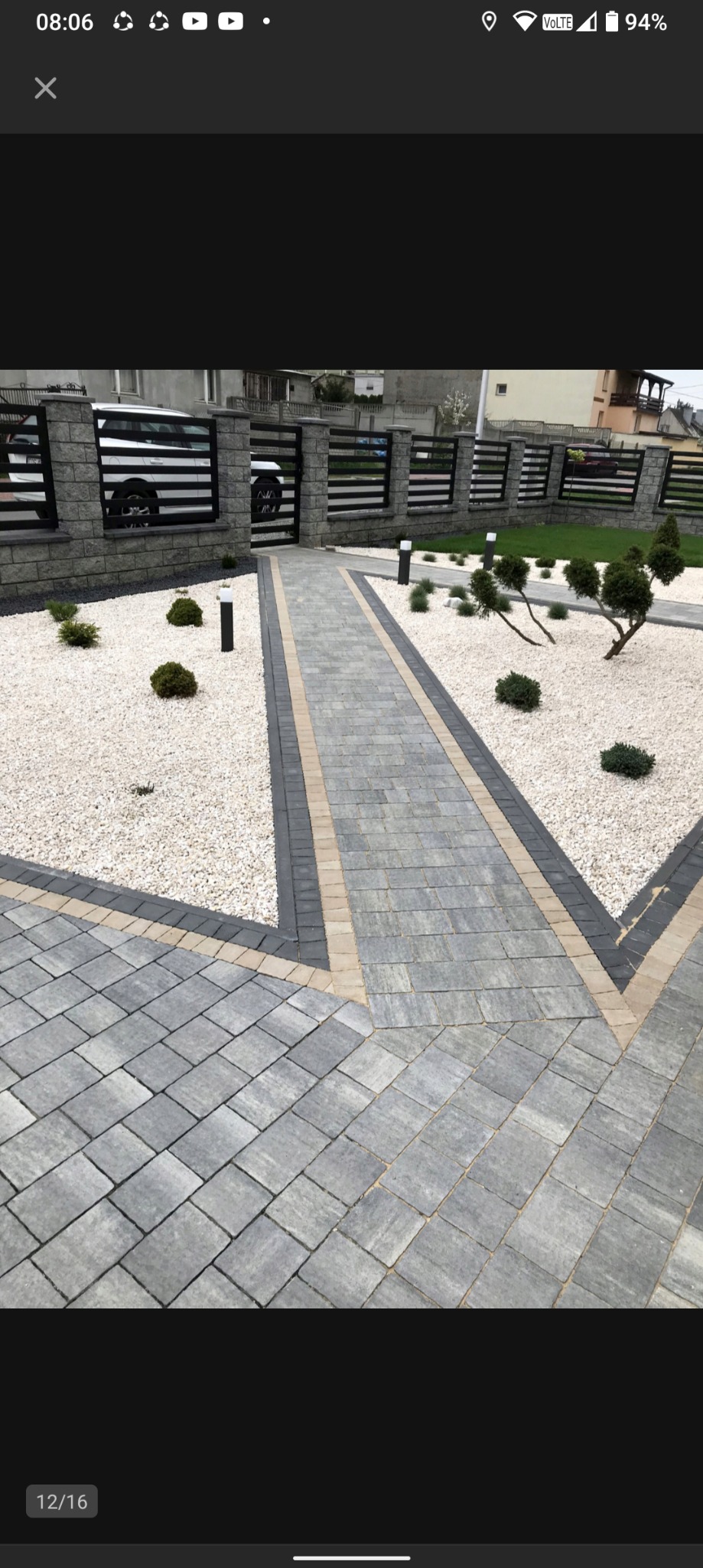 Ułożona kostka brukowa w szarym odcieniu z jasnymi kamykami i niskimi krzewami. Widoczne ogrodzenie z elementami kamiennymi. Estetyczne wykończenie podwórza.