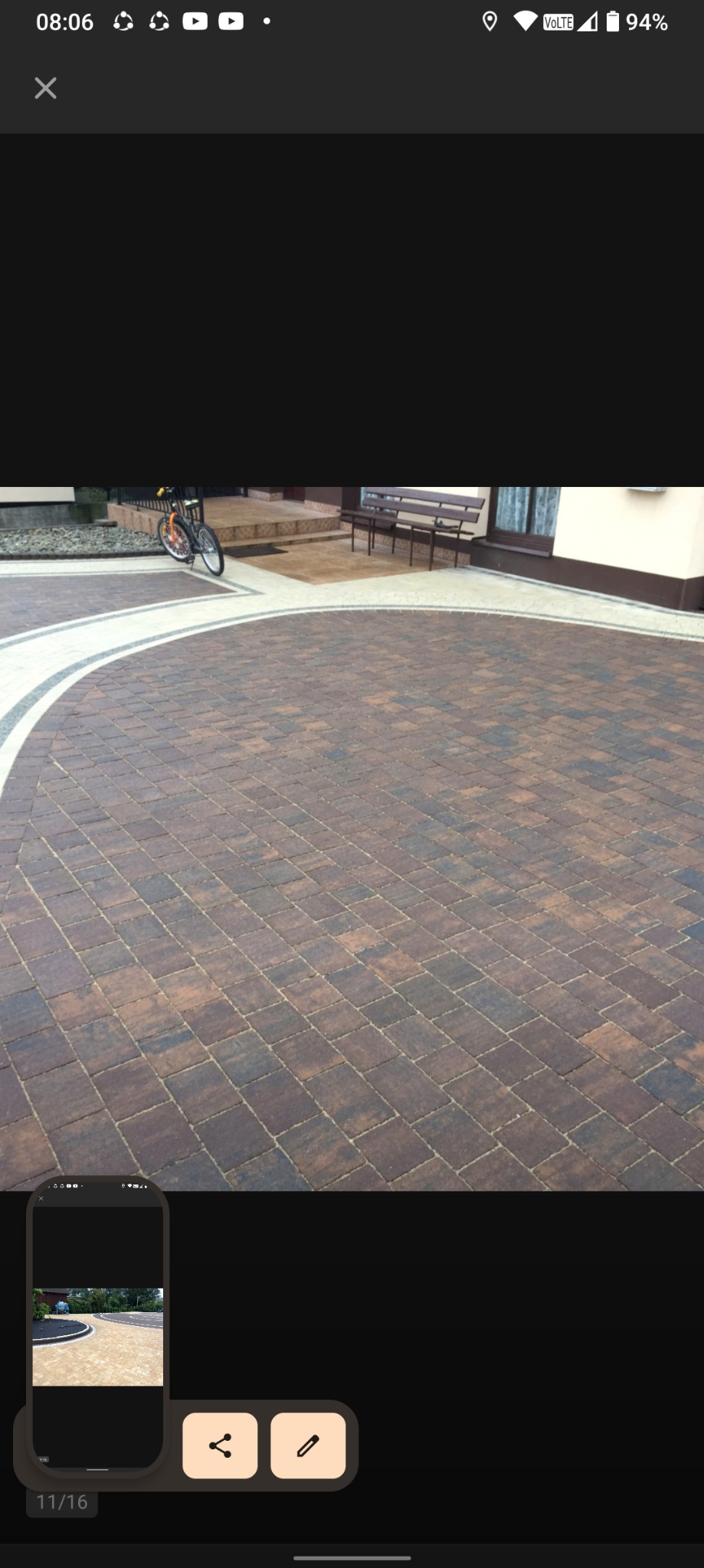 Brązowa kostka brukowa ułożona w łukowaty wzór, obrzeża z jasnej kostki, rower oparty o ścianę domu w tle, widok z perspektywy.