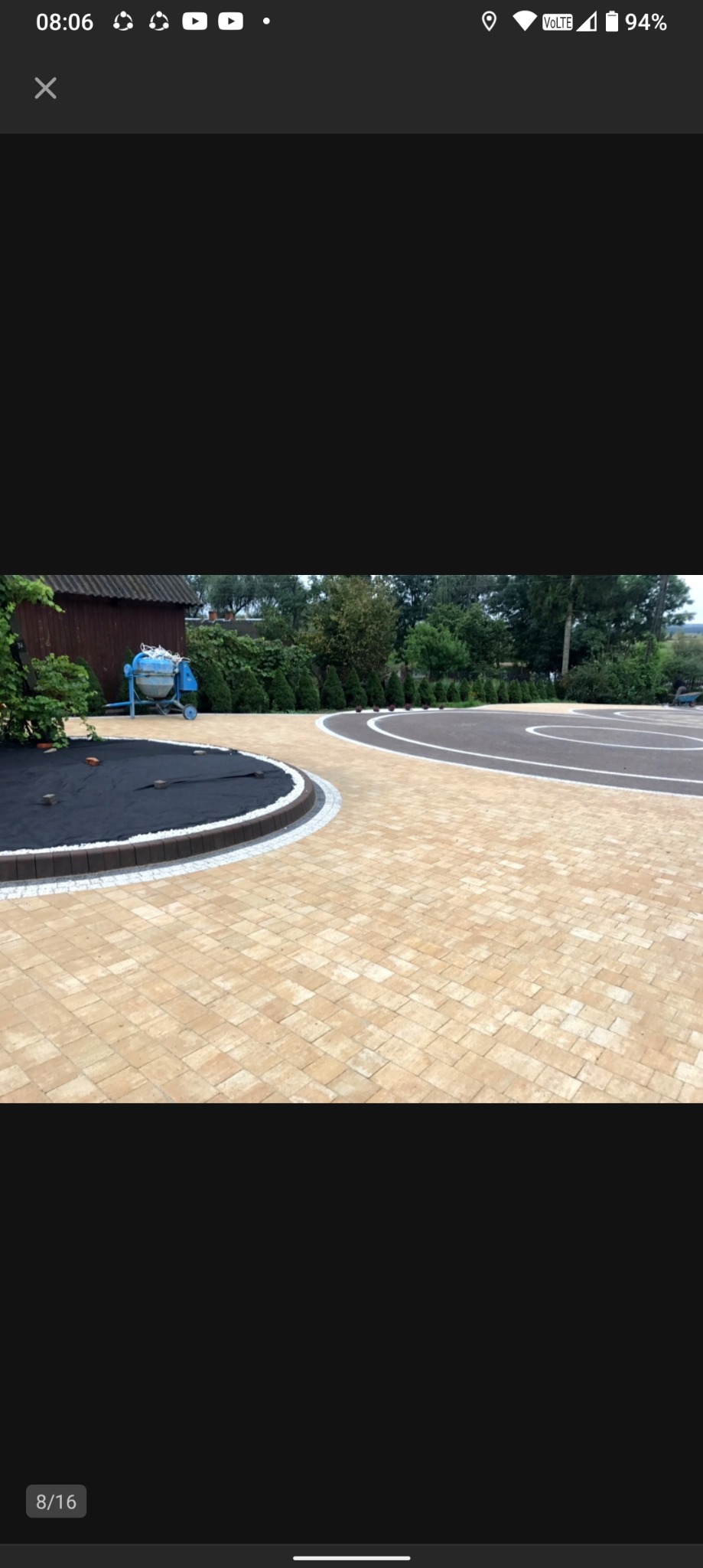 Jasna kostka brukowa ułożona w łukowaty wzór z obrzeżami. W tle widoczna zieleń i budynek gospodarczy oraz betoniarka.