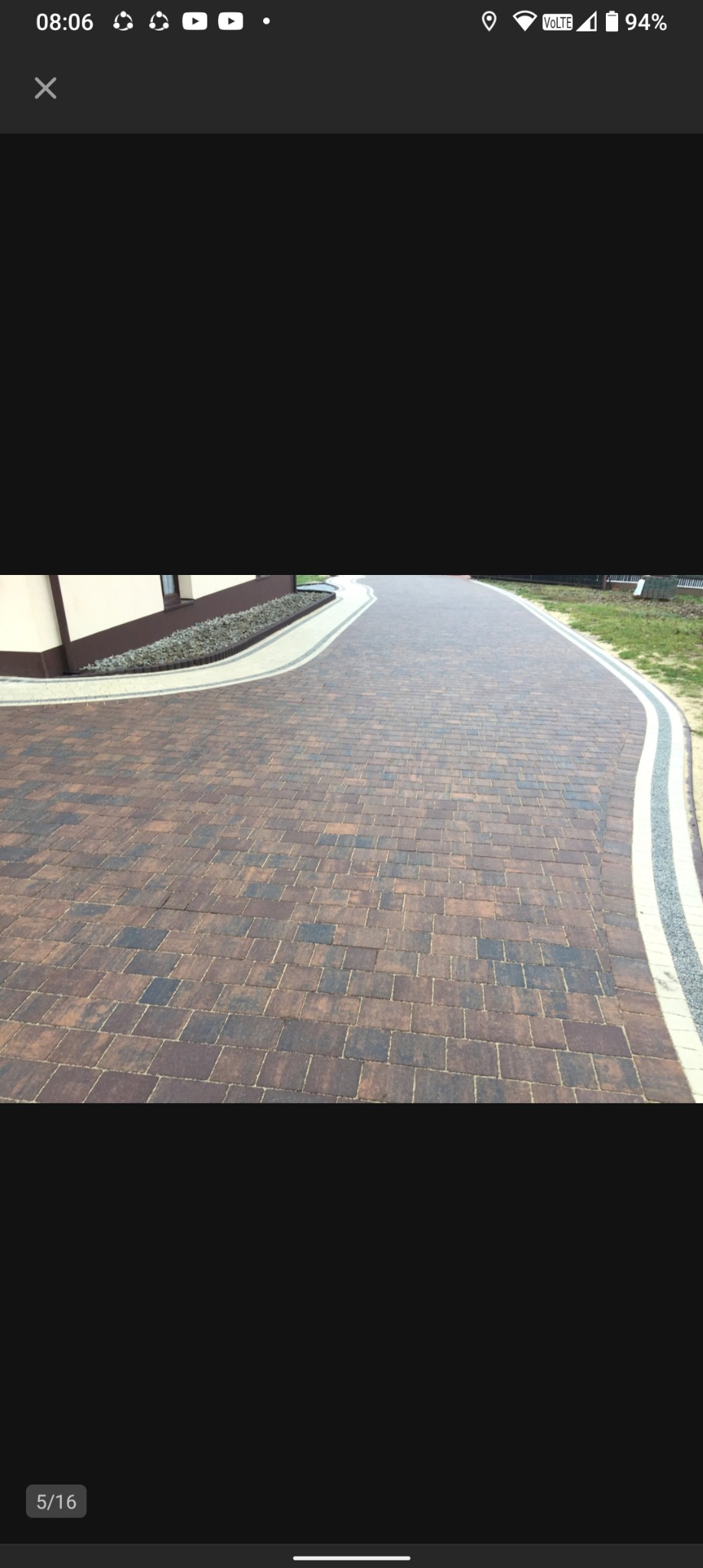 Ścieżka z brązowej kostki brukowej ułożona w łuk, obrzeża z jasnej kostki, widok z góry, w tle budynek z brązowymi akcentami i rabata z kamieniami.