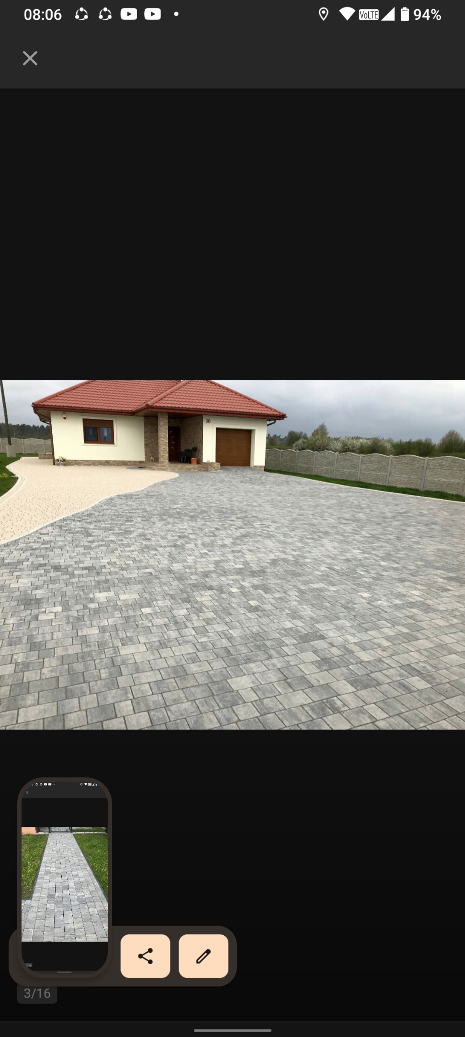 Szeroki podjazd z szarej kostki brukowej prowadzący do domu z beżową elewacją i czerwoną dachówką, oddzielony od trawnika jasnym żwirem, ogrodzony betonowym płotem.