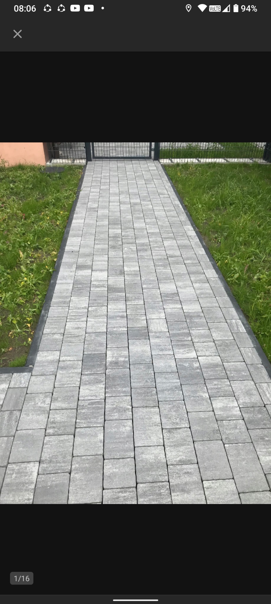 Ścieżka z szarej kostki brukowej prowadząca do metalowej bramki, otoczona zielonym trawnikiem. Krawężnik z ciemnego materiału oddziela kostkę od trawy.