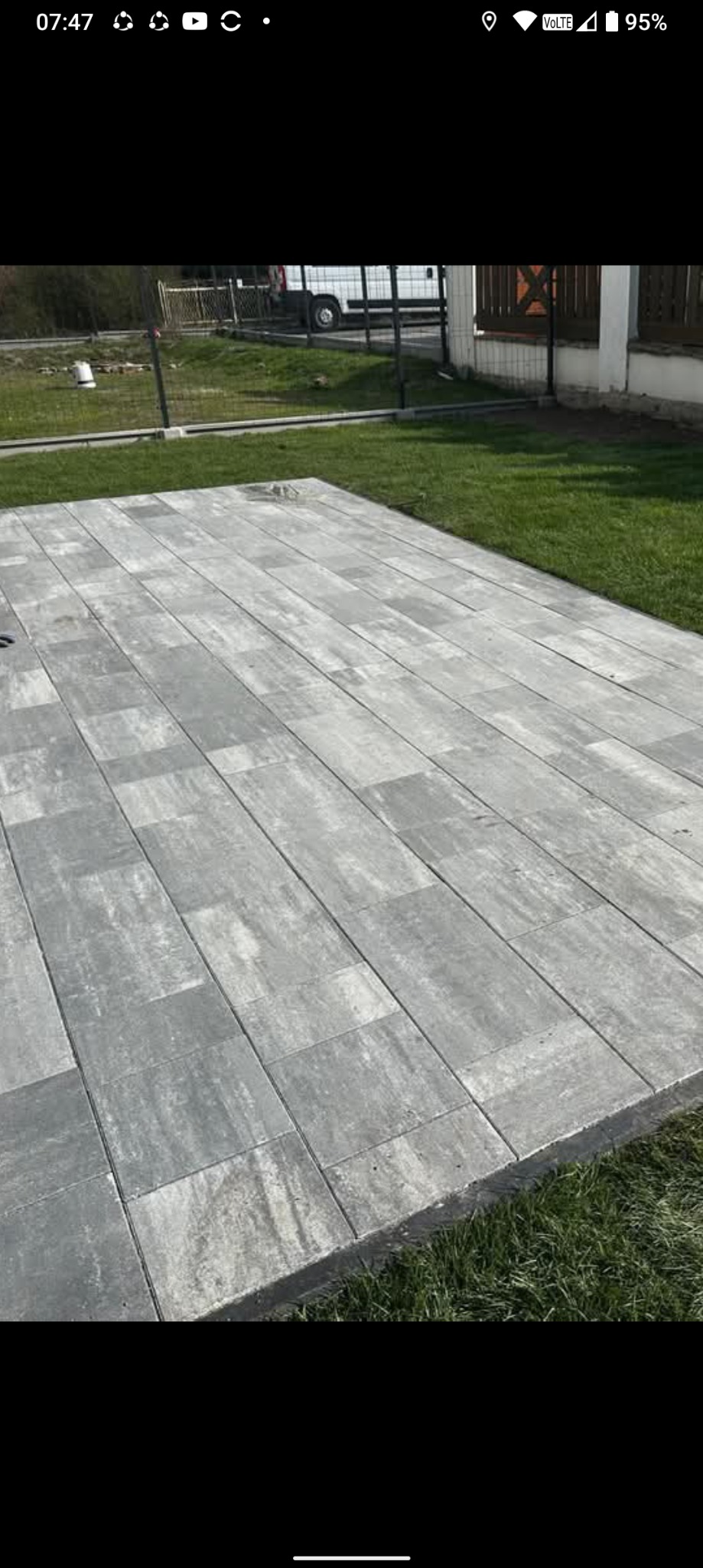 Ułożona szara kostka brukowa na trawniku. Widok z góry, regularny wzór. W tle ogrodzenie i budynek. Słoneczny dzień.