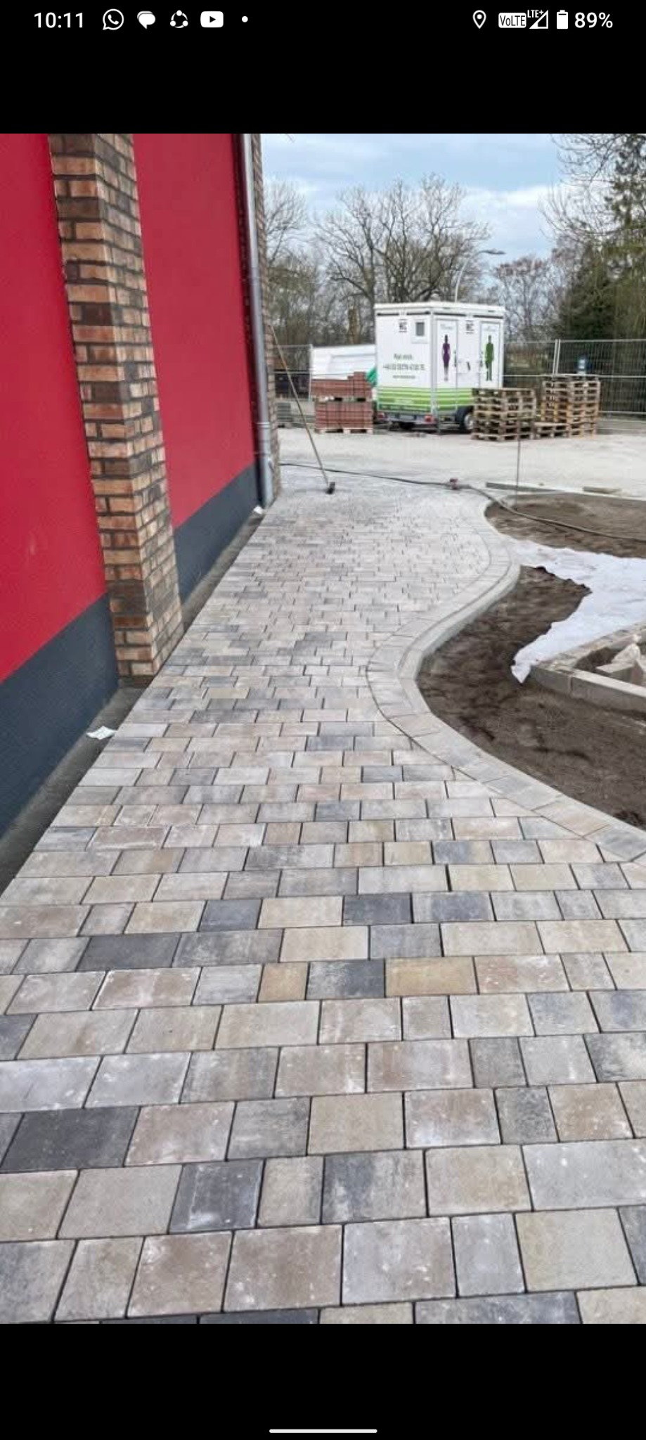 Kaskadowa ścieżka z kostki brukowej w odcieniach szarości i beżu, wijąca się obok budynku z ceglanym narożnikiem, z widocznym zapleczem budowy w tle.