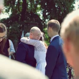 Panna młoda w koronkowej sukni i pan młody w granatowym garniturze obejmują się na tle zieleni, widoczni goście weselni.