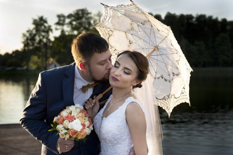Panna młoda w koronkowej sukni i welonie trzyma ozdobny parasol, całowana w policzek przez pana młodego w granatowym garniturze i muszce pod kolor bukietu ślubnego na tle jeziora.