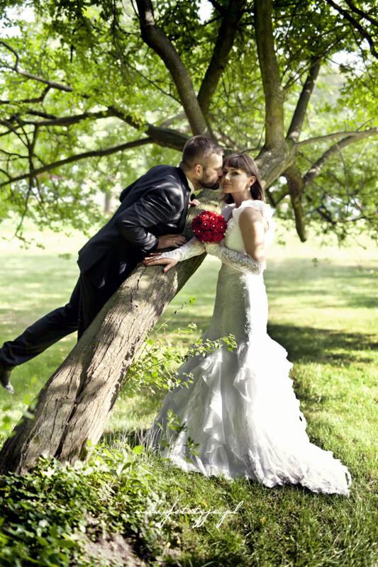 Panna młoda w koronkowej sukni i pan młody w czarnym garniturze opierają się o drzewo w parku, on całuje ją w policzek, ona trzyma bukiet czerwonych róż.