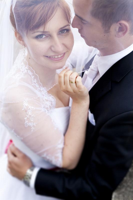 Uśmiechnięta panna młoda pod welonem, trzymająca w palcach delikatny naszyjnik, w objęciach pana młodego w czarnym garniturze. Widoczny złoty pierścionek na palcu panny młodej.