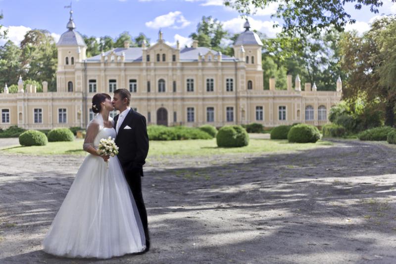 Pocałunek pary młodej na tle klasycystycznego pałacu z wieżyczkami, otoczonego zielenią i krzewami.
