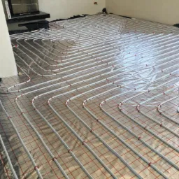 Białe rury ogrzewania podłogowego ułożone na folii z czerwoną siatką w surowym wnętrzu, widoczny kominek i okno.