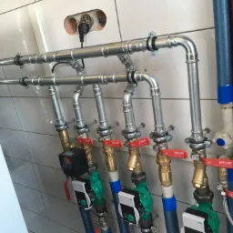 Rozbudowany system rur hydraulicznych i grzewczych z zaworami i pompami obiegowymi Wilo, zamontowany na tle białych płytek ceramicznych, widoczne także przewody elektryczne wychodzące ze ściany.