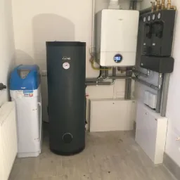 Zestaw urządzeń grzewczych w kotłowni: biały piec gazowy Bosch z wyświetlaczem, zasobnik Kospel z manometrem, filtry do wody, szara armatura i białe szafki na tle płytek podłogowych imitujących...