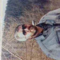 Mężczyzna w okularach przeciwsłonecznych i kurtce jeansowej leżący na ziemi porośniętej suchą trawą.