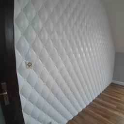 ARGIPS Arkadiusz Książek - Biała, pikowana ściana z panelami dekoracyjnymi w romby, z widocznym gniazdkiem i fragmentem drewnianej podłogi oraz ciemnej futryny drzwi.