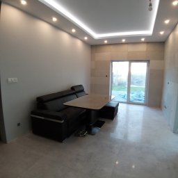 ARGIPS Arkadiusz Książek - Wykończone wnętrze salonu z szarą podłogą, ścianami i sufitem z oświetleniem LED. Widoczny czarny narożnik i stół. Przesuwne drzwi balkonowe w tle.