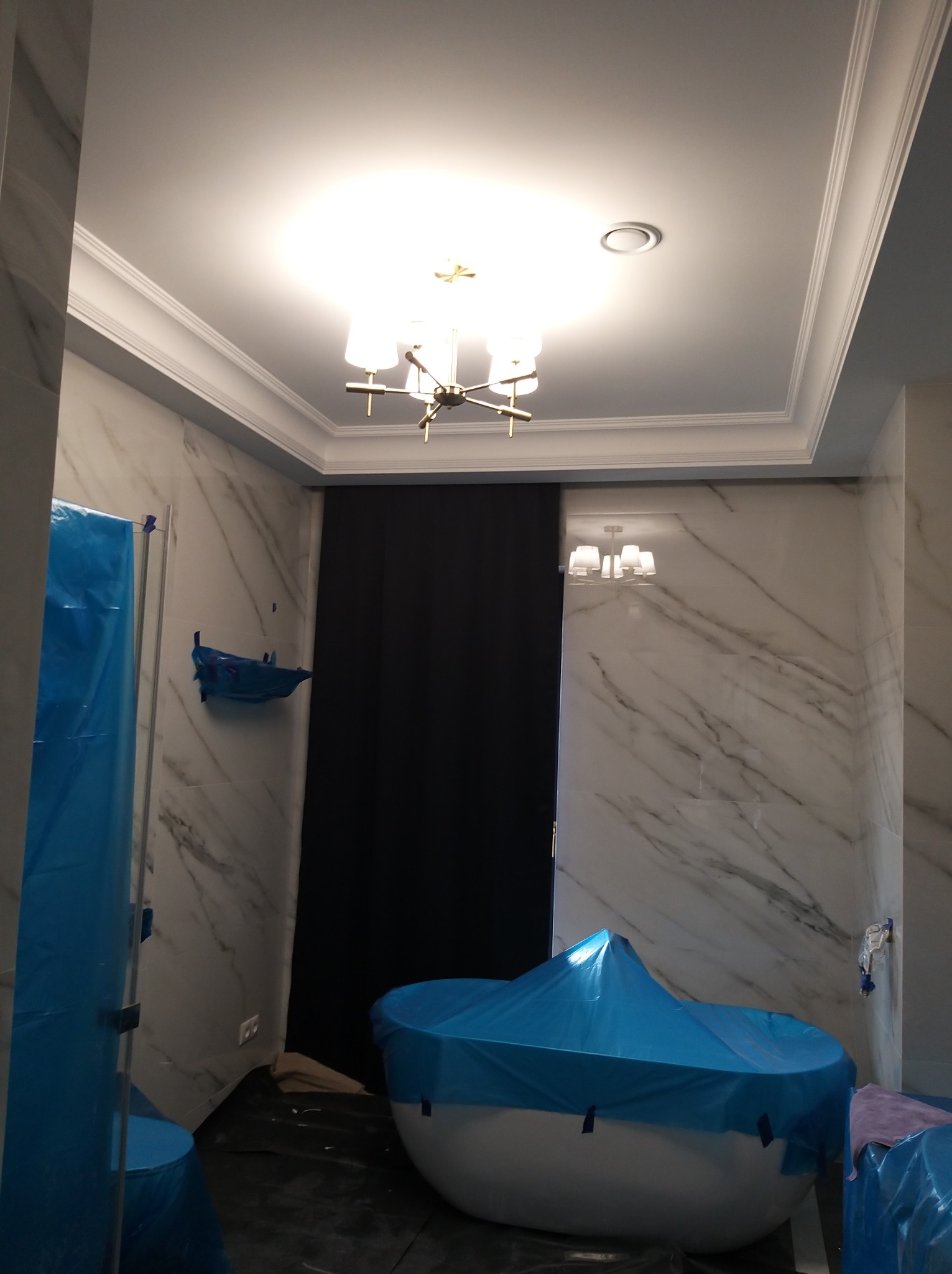 Łazienka w trakcie remontu: marmurowe płytki, zabezpieczona folią wanna i kabina prysznicowa, sufit z dekoracyjną sztukaterią i nowoczesnym oświetleniem.