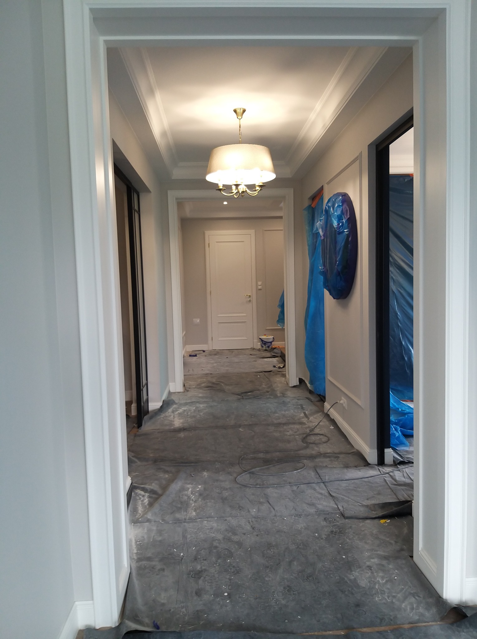 Korytarz w trakcie prac wykończeniowych: zabezpieczona podłoga, pomalowane ściany z dekoracyjnymi listwami, widoczny fragment lampy i drzwi.