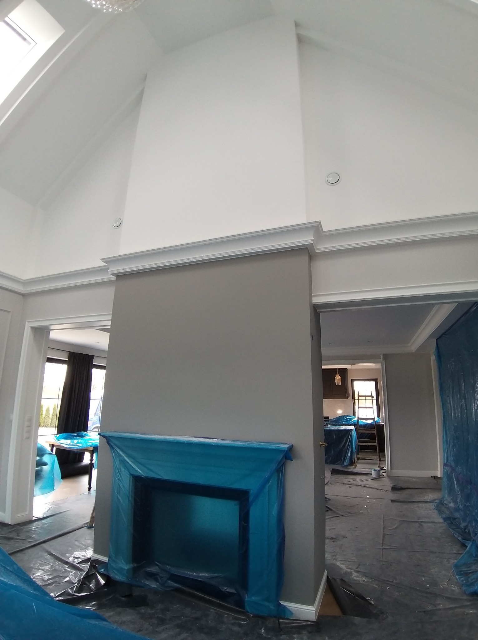 Wnętrze w trakcie malowania: szara ściana z kominkiem zabezpieczonym folią, białe listwy przysufitowe i sufit. Widoczne zabezpieczenia podłogi i mebli folią malarską.