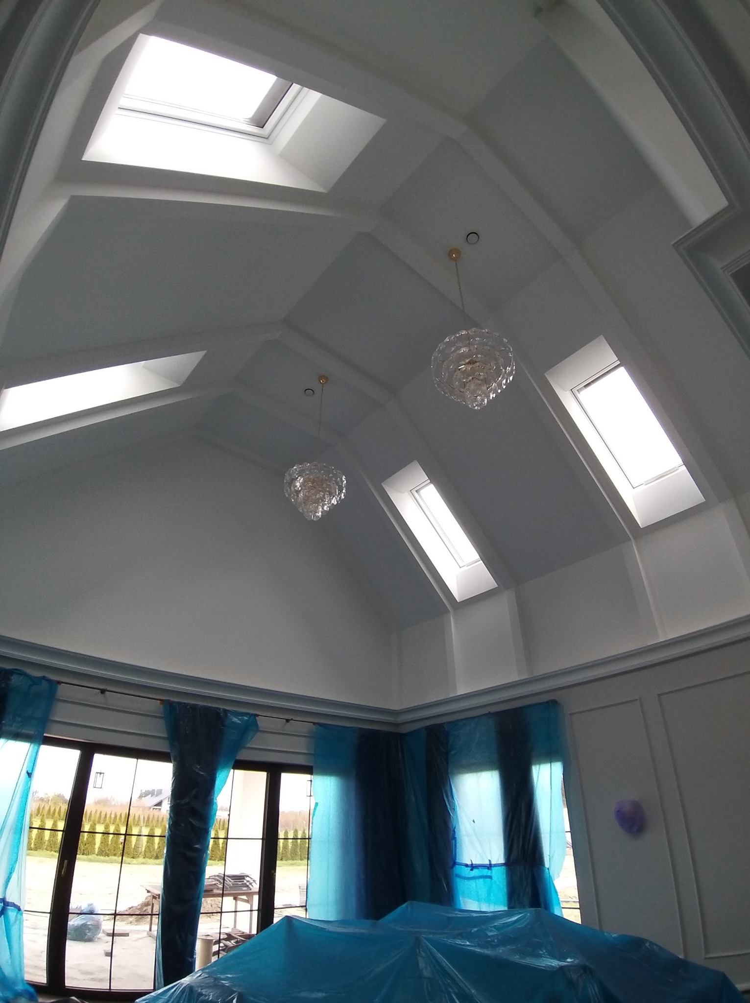 Wnętrze z wysokim skośnym sufitem i oknami dachowymi, zabezpieczone folią malarską. Widoczne dwa kryształowe żyrandole i dekoracyjne listwy ścienne.