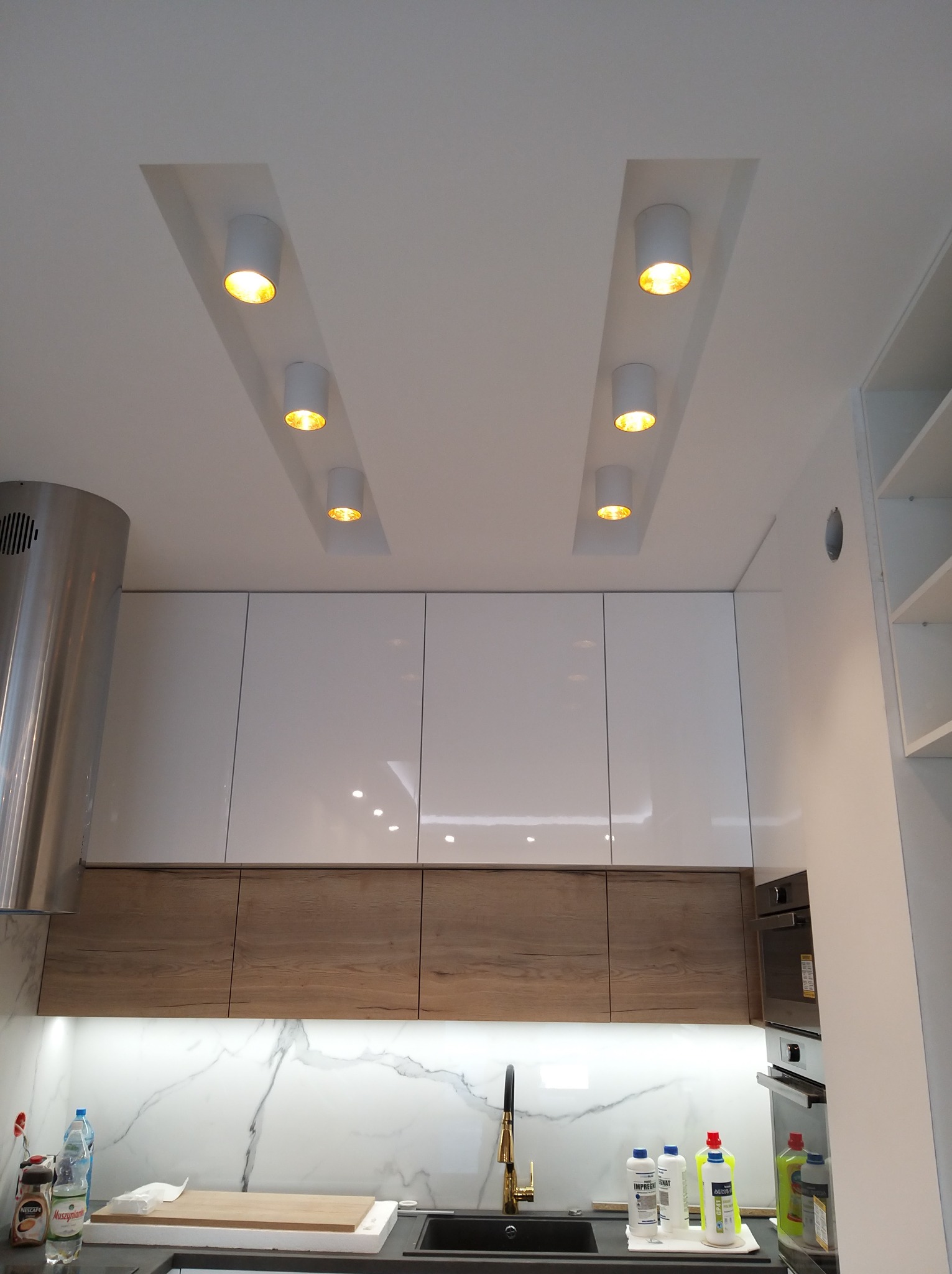 Nowoczesna kuchnia z białymi szafkami, drewnianymi frontami i marmurowym backsplashiem. Sufit z wbudowanymi, okrągłymi lampami punktowymi. Minimalistyczny design.